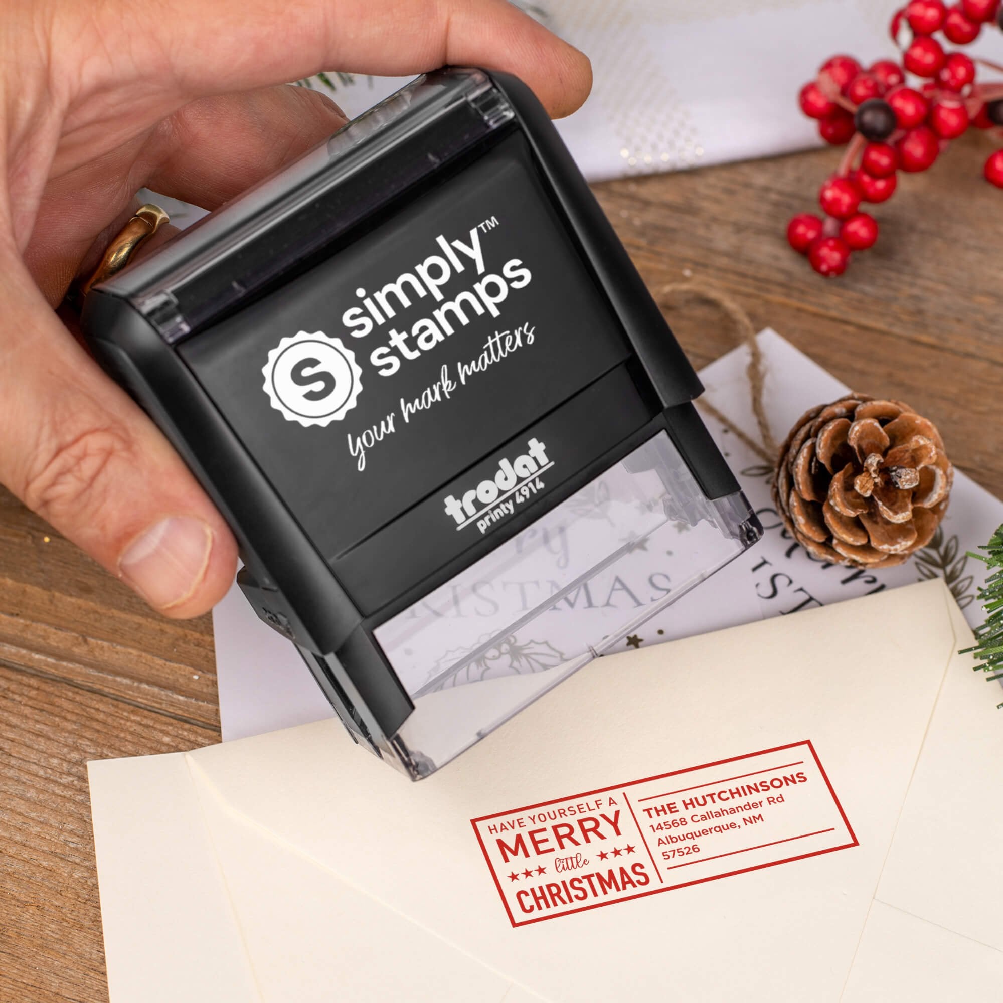 Holiday-themed ‘Have Yourself A Merry Little Christmas’ address stamp, hand using self-inking Christmas tree address stamp to personalize a holiday envelope on festive wooden table.